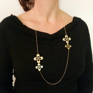 May include: A gold chain necklace with three flower-shaped pendants. The necklace is on a model wearing a black shirt.