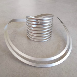 Pu&ograve; includere: Un anello in metallo argentato composto da pi&ugrave; bande sottili intrecciate insieme per formare una forma circolare. L'anello &egrave; su una superficie grigio chiaro.