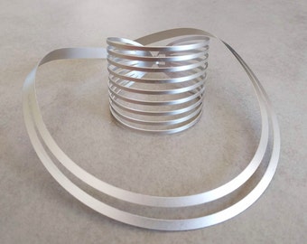 Puede incluir: Un anillo de metal plateado hecho de múltiples bandas delgadas retorcidas juntas para formar una forma circular. El anillo está sobre una superficie gris claro.