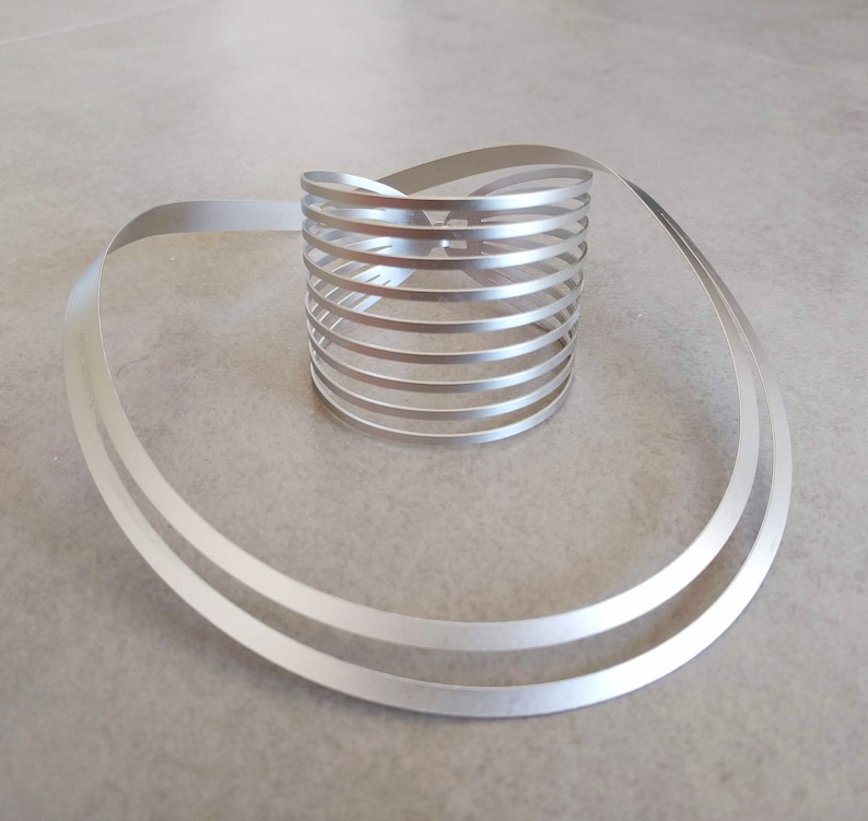 Pu&ograve; includere: Un anello in metallo argentato composto da pi&ugrave; bande sottili intrecciate insieme per formare una forma circolare. L'anello &egrave; su una superficie grigio chiaro.