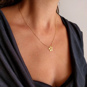 May include: A gold chain necklace with a delicate star pendant. The necklace is worn by a person with dark hair and a black shirt.
