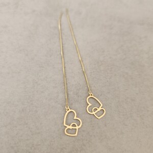 Può includere: Orecchini pendenti color oro con un design a doppio cuore. Gli orecchini hanno una delicata catena che pende, con un ciondolo a forma di cuore all'estremità. Gli orecchini sono esposti su una superficie grigia.