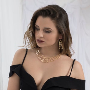 May include: A woman wearing a black off-the-shoulder dress with a gold geometric necklace and matching earrings.