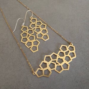 Pu&ograve; includere: Una collana e orecchini abbinati in oro con un design geometrico a nido d'ape. La collana &egrave; una catena delicata con un ciondolo che &egrave; una serie di forme esagonali collegate. Gli orecchini sono anche esagonali e pendono da piccoli ganci dorati.