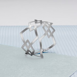 Puede incluir: Un brazalete geométrico plateado con un patrón de diamante repetido. El brazalete está hecho de metal y tiene un acabado pulido.