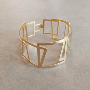 Può includere: Un braccialetto in metallo dorato con un design geometrico. Il braccialetto è realizzato con sottili strisce di metallo che sono collegate per formare una serie di quadrati e triangoli.