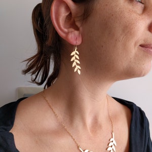 May include: A gold necklace and earrings featuring a delicate leaf design. The necklace is a simple chain with a pendant of leaves arranged in a curved shape. The earrings are long and dangle, with leaves arranged in a vertical line.