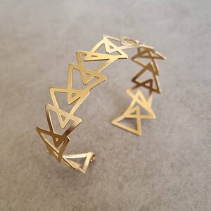 May include: A gold-toned metal bracelet with a geometric design featuring a series of interlocking triangles.