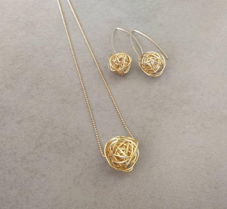 May include: A gold necklace and earrings set with a delicate chain and a round, wire-wrapped pendant. The pendant and earrings are made of a woven wire design.