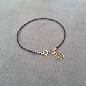 May include: A black string bracelet with a gold hamsa charm. The hamsa charm has a detailed design with lines.