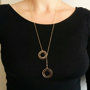 May include: A gold necklace with a long chain and two pendant circles. The circles are made of multiple concentric rings.