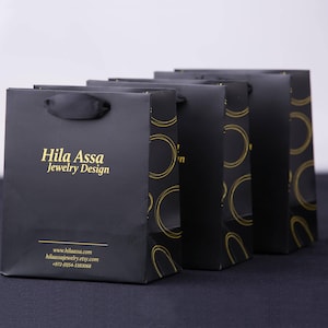 Puede incluir: Cinco bolsas de papel negras con patrones circulares dorados y una cinta negra. La bolsa de la izquierda tiene el texto "Hila Assa Jewelry Design" en letras doradas y la direcci&oacute;n del sitio web "www.hilaassa.com" y "hilaassa.jewelry.etsy.com" en letras doradas m&aacute;s peque&ntilde;as.
