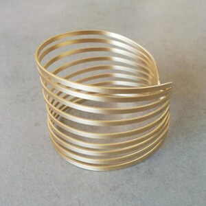 May include: A gold-toned metal bangle bracelet with a multi-layered design. The bracelet is made of several thin, overlapping bands that create a unique and stylish look.