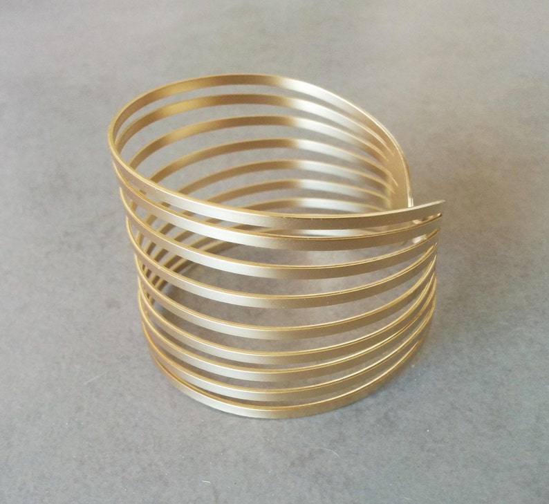 May include: A gold-toned metal bangle bracelet with a multi-layered design. The bracelet is made of several thin, overlapping bands that create a unique and stylish look.