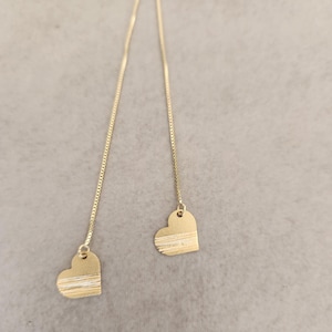 Può includere: Orecchini pendenti color oro con ciondoli a forma di cuore. Ogni cuore ha una finitura strutturata e spazzolata con linee orizzontali. Gli orecchini pendono da delicate catene su uno sfondo neutro.