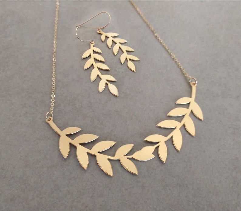 May include: A gold necklace and earrings set featuring a delicate leaf design. The necklace is a single strand with a delicate chain and a pendant shaped like a branch with leaves. The earrings are simple studs with a leaf design.