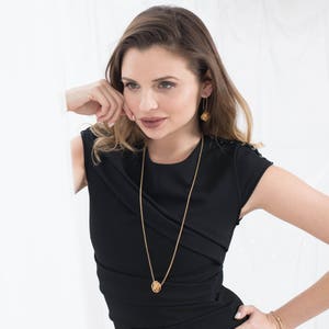 May include: A woman wearing a black dress with a gold chain necklace and gold earrings. The necklace features a small, round pendant.