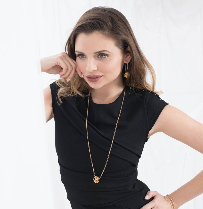 May include: A woman wearing a black dress with a gold chain necklace and gold earrings. The necklace features a small, round pendant.