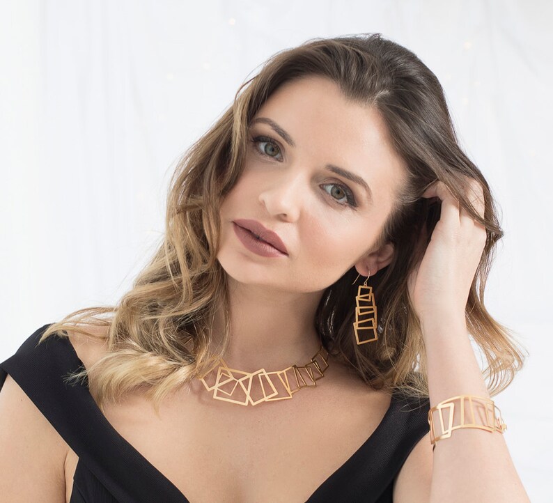 May include: A gold geometric necklace and earrings set with a modern, abstract design. The necklace features a series of interconnected squares and rectangles, while the earrings are long and dangle, with stacked squares.