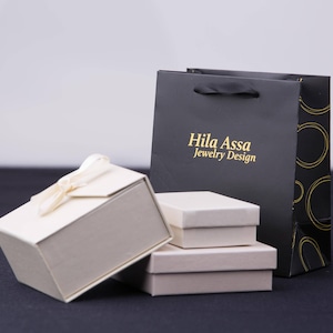 Puede incluir: Un conjunto de tres cajas de regalo blancas con una bolsa de compras negra y dorada. La bolsa de compras tiene el texto "Hila Assa Jewelry Design" impreso en ella.