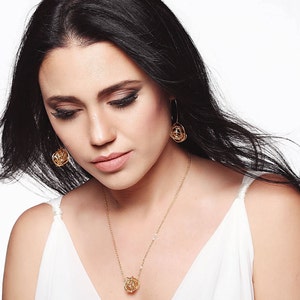 May include: A woman wearing a white top with a gold necklace and matching earrings. The necklace and earrings feature a delicate, intricate design.