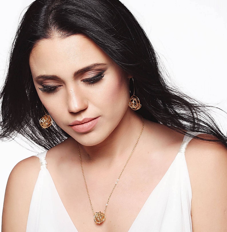 May include: A woman wearing a white top with a gold necklace and matching earrings. The necklace and earrings feature a delicate, intricate design.