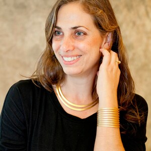 Op de afbeelding: Een vrouw met lang bruin haar draagt een zwart shirt met een gouden ketting en een gouden armband. Ze glimlacht en kijkt naar de zijkant.