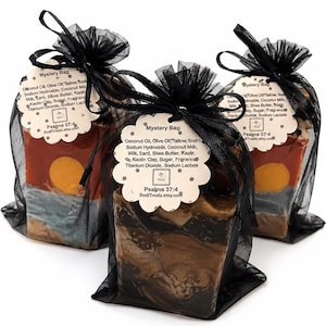 May include: Three rectangular bars of soap packaged in black organza drawstring bags. Each bag features a scalloped tag labeled Mystery Bag with ingredients including coconut oil, olive oil, tallow, and shea butter, plus the text Psalms 37:4.