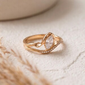 Puede incluir: Un anillo de oro con una piedra preciosa transparente central de talla marquesa en engaste de garras, con una banda curva que incorpora una hilera delicada de piedras transparentes en pavé a lo largo de un lado del aro.