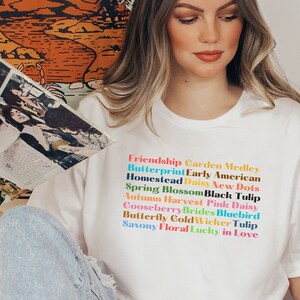 May include: White t-shirt with a colorful text design that reads "Friendship Garden Medley, Butterprint Early American, Homestead Daisy New Dots, Spring Blossom Black Tulip, Autumn Harvest Pink Daisy, Gooseberry Brides Bluebird, Butterfly Cold Wicker Tulip, Saxony Floral Lucky in Love."