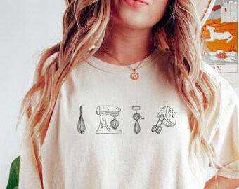May include: A cream-colored t-shirt with a graphic of kitchen tools, including a whisk, a stand mixer, and a hand mixer.