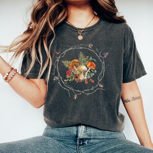 May include: A dark gray t-shirt with a graphic of mushrooms, ferns, and acorns in a wreath design. The text "More Getaway" is tattooed on the wearer's arm.