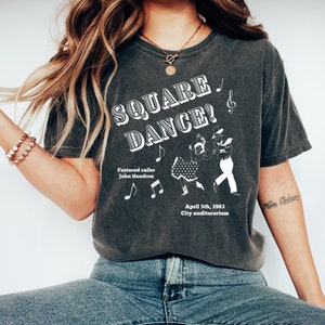May include: A dark grey t-shirt with a white graphic design featuring a square dance scene. The text on the shirt reads "SQUARE DANCE!" with the words "Featured caller John Hendron" and "April 5th, 1963 City auditorium" below the graphic.