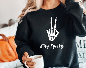 Może przedstawiać: Czarny sweter z białą ręką szkieletu robiącą znak pokoju i tekstem "Stay Spooky".