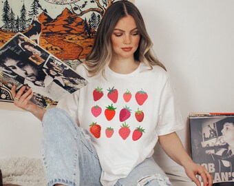 Puede incluir: Una camiseta blanca con un patrón de fresas rojas. La camiseta la lleva puesta una persona con el pelo largo y castaño.