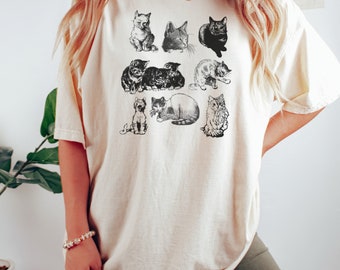 May include: A cream-colored t-shirt with a graphic print of nine black and white cats. The cats are in various poses, including sitting, lying down, and standing. The shirt is oversized and has a relaxed fit.