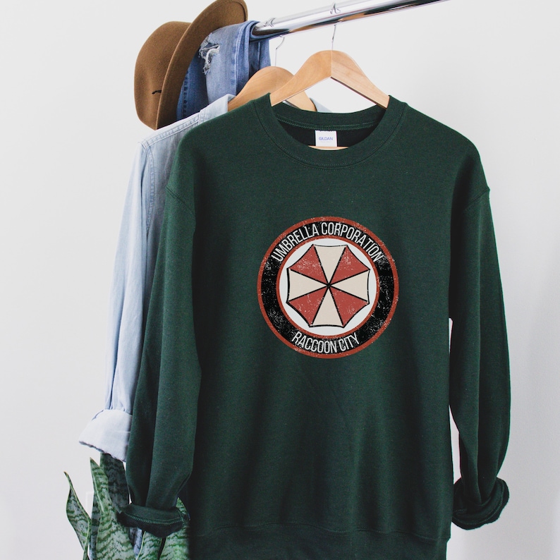 May include: A dark green sweatshirt with a vintage-style logo featuring a red and white umbrella with the text "Umbrella Corporation Raccoon City".