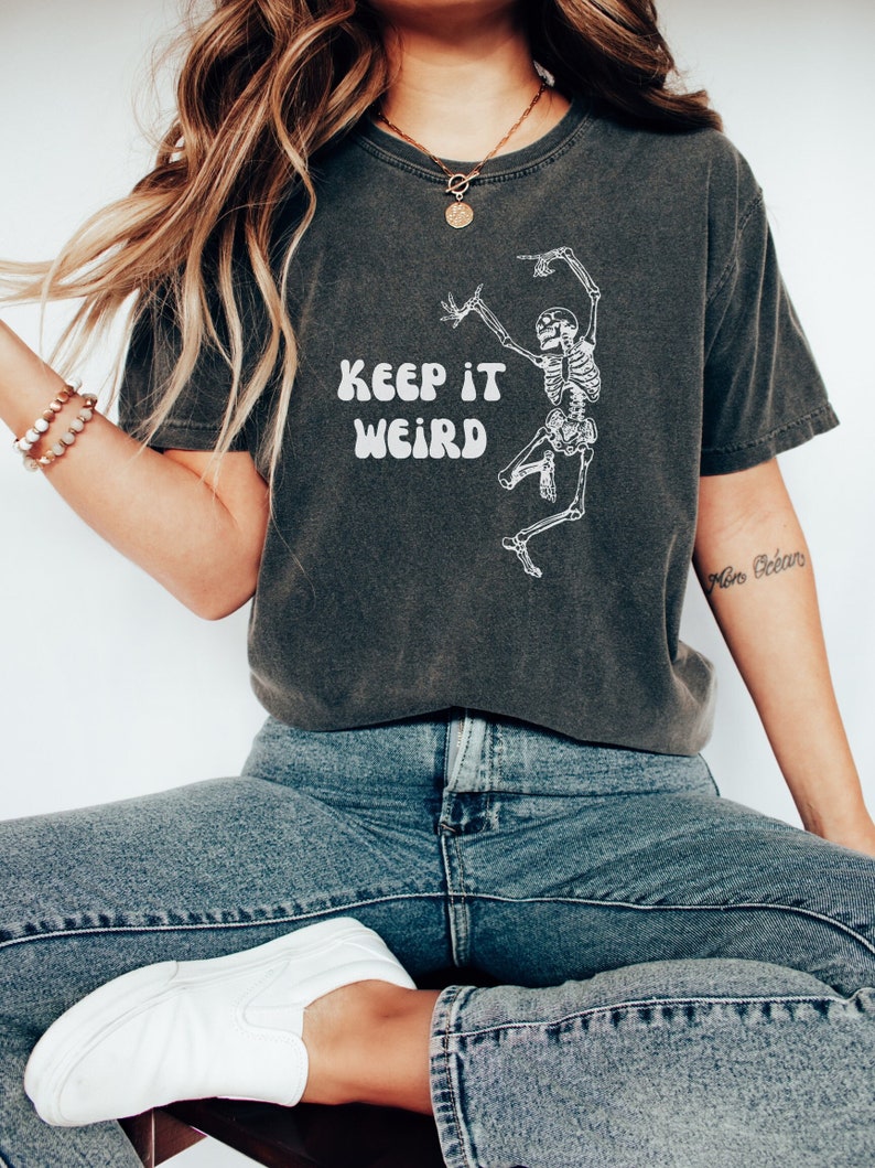 May include: A dark gray t-shirt with a white skeleton dancing and the text "Keep it Weird" in a retro font.