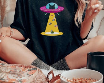 May include: A black t-shirt with a graphic of a black cat in a flying saucer. The saucer is pink and purple with yellow lights. The cat is looking down at a pile of cat food cans.