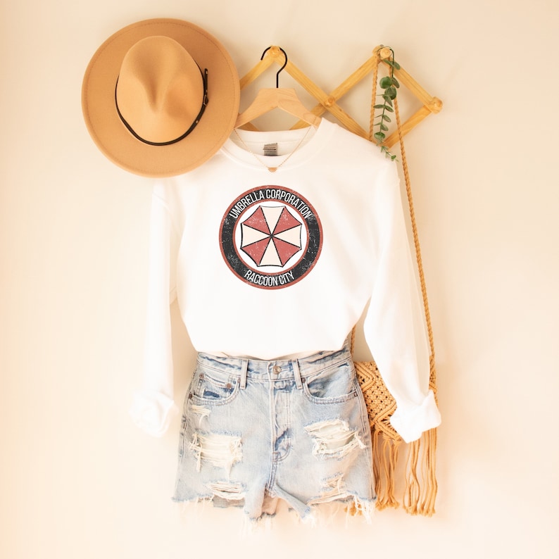 May include: White long-sleeved sweatshirt with a red and white Umbrella Corporation logo on the front. The logo is a black circle with the words "Umbrella Corporation" and "Raccoon City" in white text. The sweatshirt is hanging on a wooden hanger.