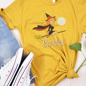 Op de afbeelding: Een geel T-shirt met een afbeelding van een heks die op een bezem vliegt. De heks draagt een zwarte jurk en een punthoed. De tekst "Bewitched" is onder de afbeelding gedrukt.