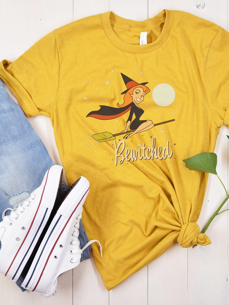 Op de afbeelding: Een geel T-shirt met een afbeelding van een heks die op een bezem vliegt. De heks draagt een zwarte jurk en een punthoed. De tekst "Bewitched" is onder de afbeelding gedrukt.