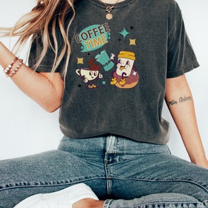Op de afbeelding: Een donkergrijs T-shirt met een cartoon afbeelding van een koffiekopje en een donut. De tekst "Coffee Time!" staat op het shirt gedrukt.