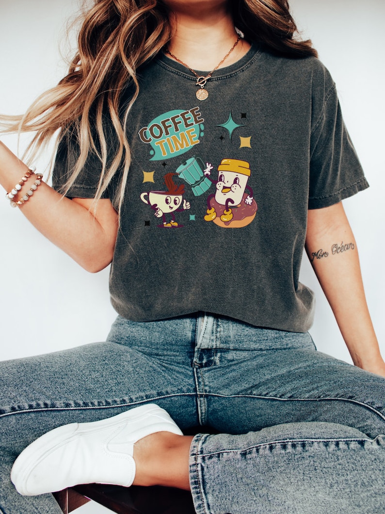 Op de afbeelding: Een donkergrijs T-shirt met een cartoon afbeelding van een koffiekopje en een donut. De tekst "Coffee Time!" staat op het shirt gedrukt.