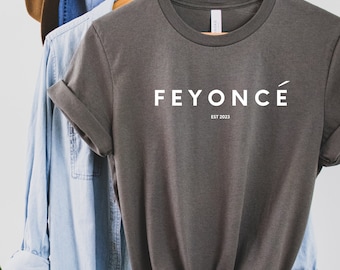 Può includere: Una maglietta verde scuro con la scritta "FEYONCÉ EST 2023" stampata in bianco sul davanti.