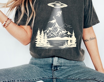 Könnte beinhalten: Ein dunkelgraues T-Shirt mit einem Aufdruck eines UFOs, das einen Bigfoot vor einer Bergkette entführt. Der Aufdruck ist im Vintage-Stil mit einer hellgelben Farbe gehalten.