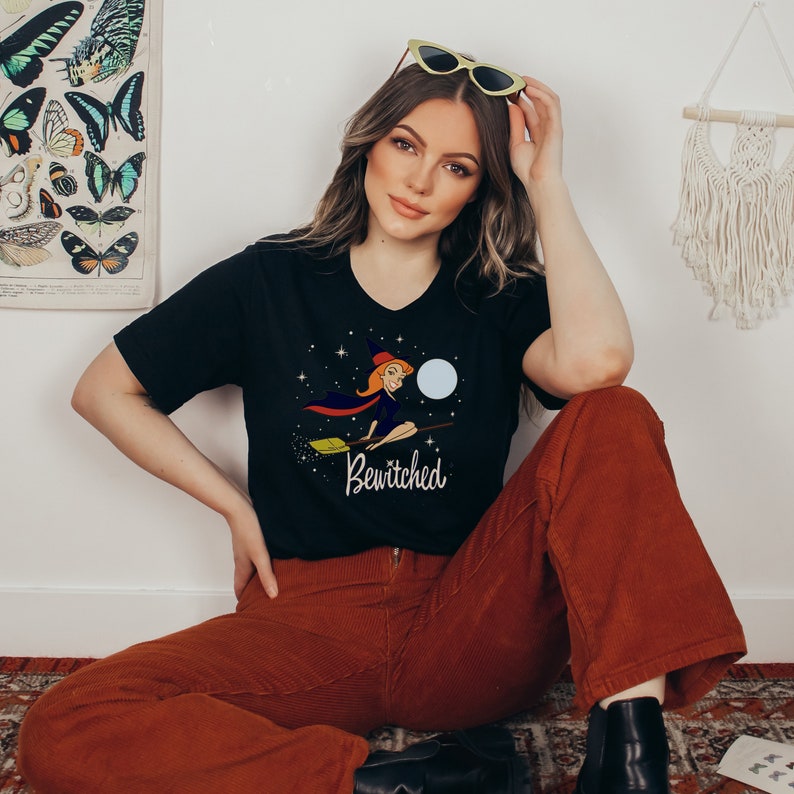 Op de afbeelding: Een zwart T-shirt met een afbeelding van een heks die op een bezem vliegt met de tekst "Bewitched" eronder. De heks draagt een zwarte jurk en een heksenhoed. De achtergrond is een sterrenhemel met een volle maan.