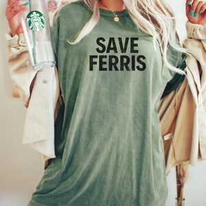 Pode incluir: Uma mulher vestindo uma camiseta verde de tamanho grande com o texto "SAVE FERRIS" estampado na frente. Ela est&aacute; segurando um copo de pl&aacute;stico transparente com uma tampa verde.