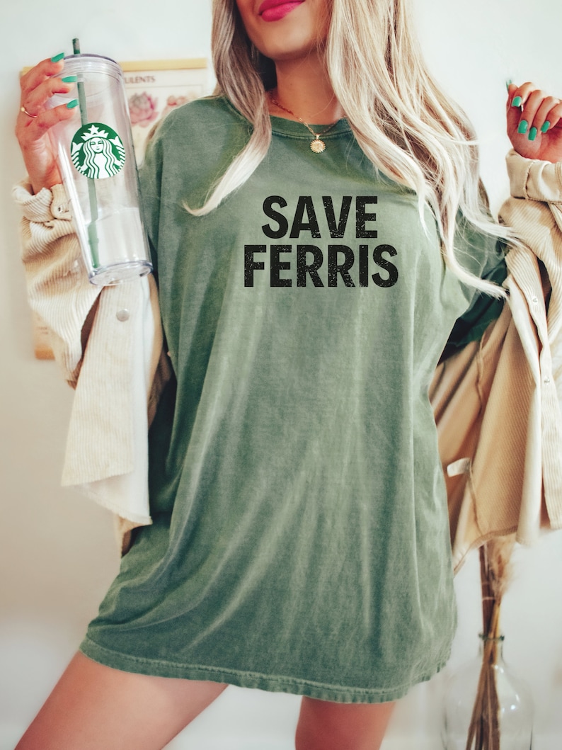 Pode incluir: Uma mulher vestindo uma camiseta verde de tamanho grande com o texto "SAVE FERRIS" estampado na frente. Ela est&aacute; segurando um copo de pl&aacute;stico transparente com uma tampa verde.