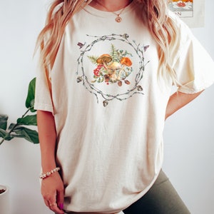 May include: A cream-colored oversized t-shirt with a graphic of mushrooms and greenery in a wreath design.